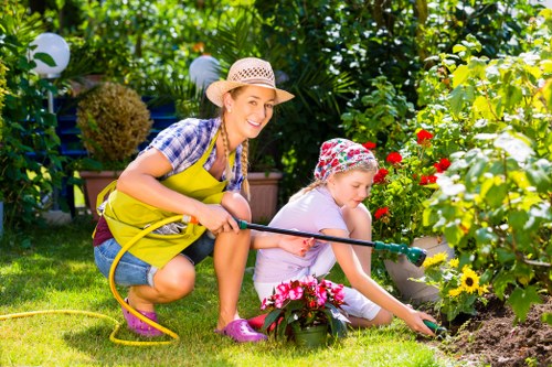 In an outdoor garden area, a woman and a young girl are engaged in gardening activities on a sunny day. The woman, wearing a straw hat, yellow apron, and casual clothing, is smiling as she holds a garden hose, suggesting she is watering plants. The girl, dressed in light clothing with a colorful hat, is kneeling on the grass near a flower bed, carefully tending to pink and yellow flowers. The garden features a well-maintained lawn with lush green grass, bordered by flower beds filled with bright, blooming flowers. In the background, mature shrubs and trees provide shade and structure to the landscaped space, along with decorative garden lighting hanging from the trees. The overall scene depicts a vibrant and tidy outdoor area in Bethnal Green, with natural sunlight illuminating the setting, highlighting the healthy plant life and the outdoor work of gardening. The scene emphasizes gardening and outdoor maintenance, consistent with services offered by Pressure Washing Bethnal Green, within a well-kept residential garden environment.